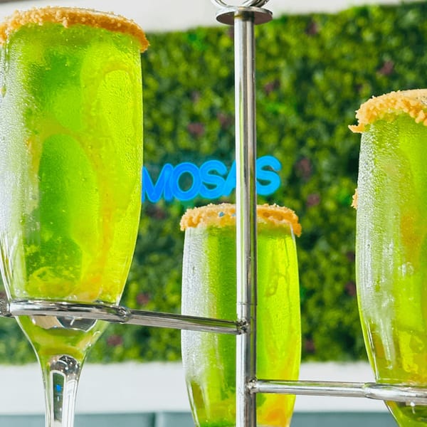 Three green mimosas with sugared rims on a metal stand, with a neon "MIMOSAS" sign in the background.