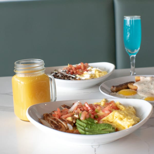 Three breakfast plates with eggs, meat, and toppings, alongside orange juice and a blue drink.
