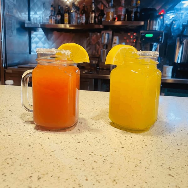 Two mason jar drinks, one orange and one yellow, garnished with lemon slices and salt.