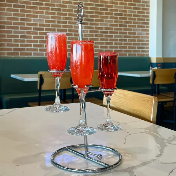 Three Easy Egg mimosas with red sugar rims on a chrome stand, against a brick wall and green booth.