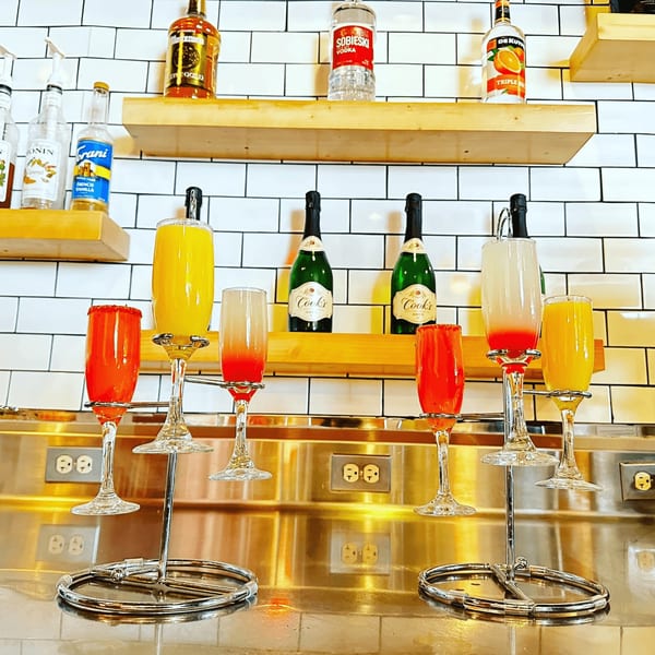 A mimosa flight with orange and red drinks, champagne bottles, and liquor bottles on shelves.