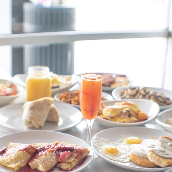 A table laden with various breakfast dishes, including crepes, eggs, and a mimosa.