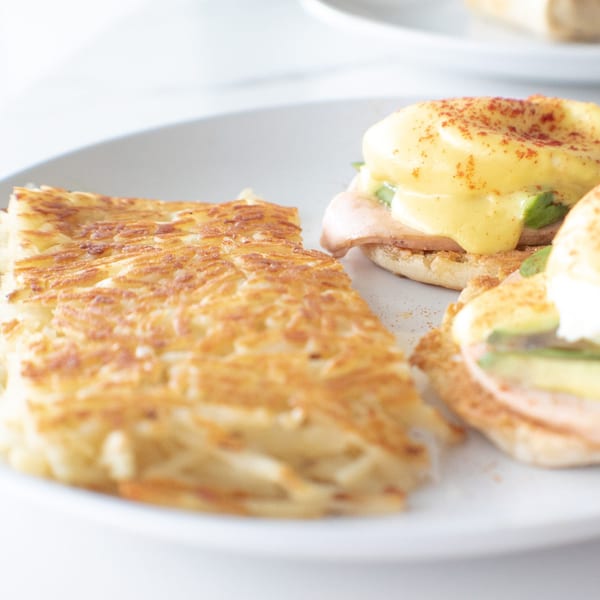 Hash browns and two Eggs Benedict with hollandaise sauce, ham, and avocado on a white plate.