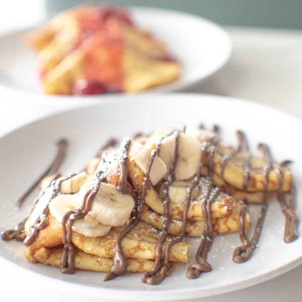 Crepes topped with sliced bananas, chocolate drizzle, and powdered sugar on a white plate.
