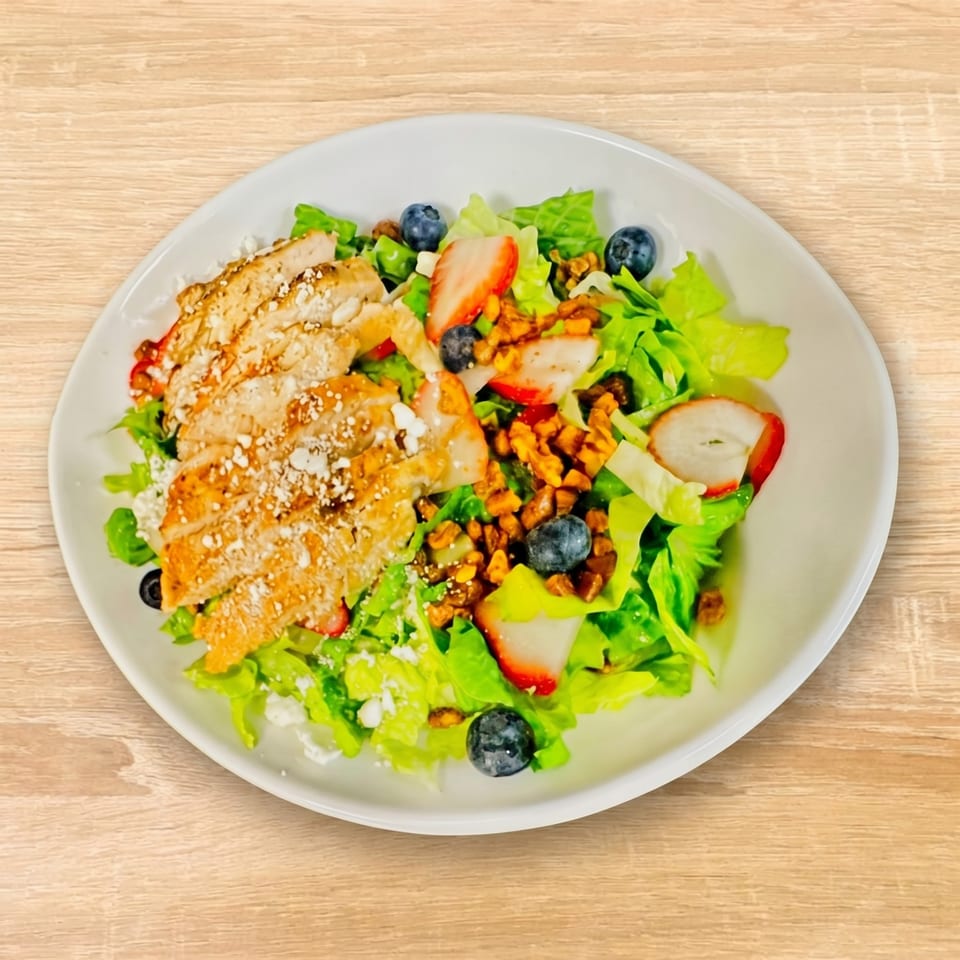Grilled chicken salad with lettuce, strawberries, blueberries, walnuts, and feta cheese on a white plate.