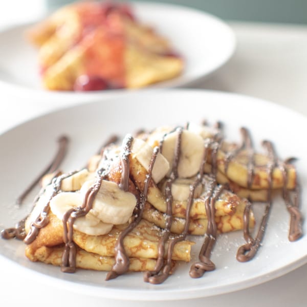 Banana and chocolate drizzled crepes with powdered sugar, with berry crepes blurred in the background.