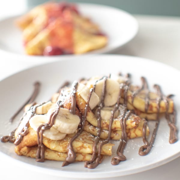 Crepes topped with sliced bananas, chocolate drizzle, and powdered sugar, with another plate in the background.