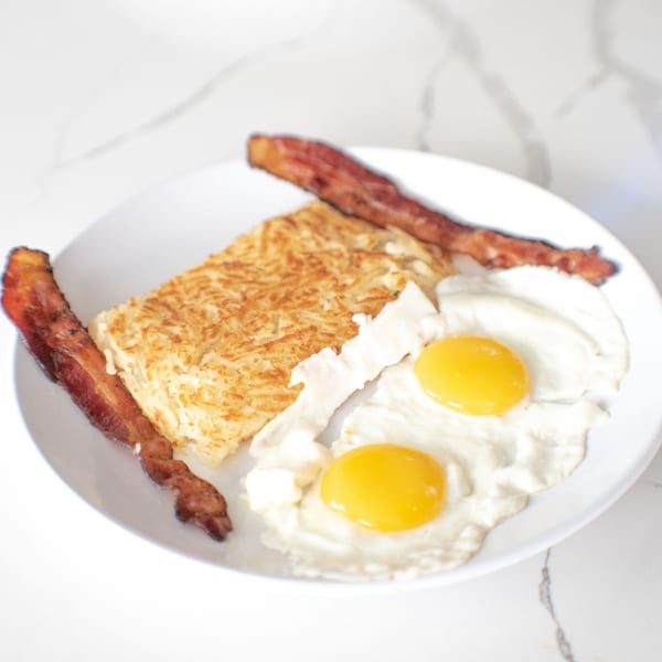 A breakfast plate with two fried eggs, a large hash brown patty, and two strips of bacon.