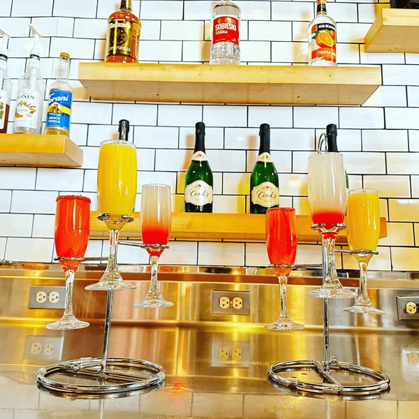 A mimosa flight with orange and red drinks, with champagne bottles and liquor on shelves.
