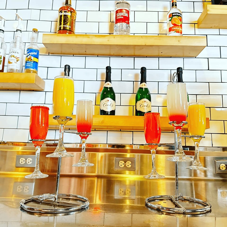 A mimosa flight with orange and red drinks, with champagne bottles and liquor on shelves.